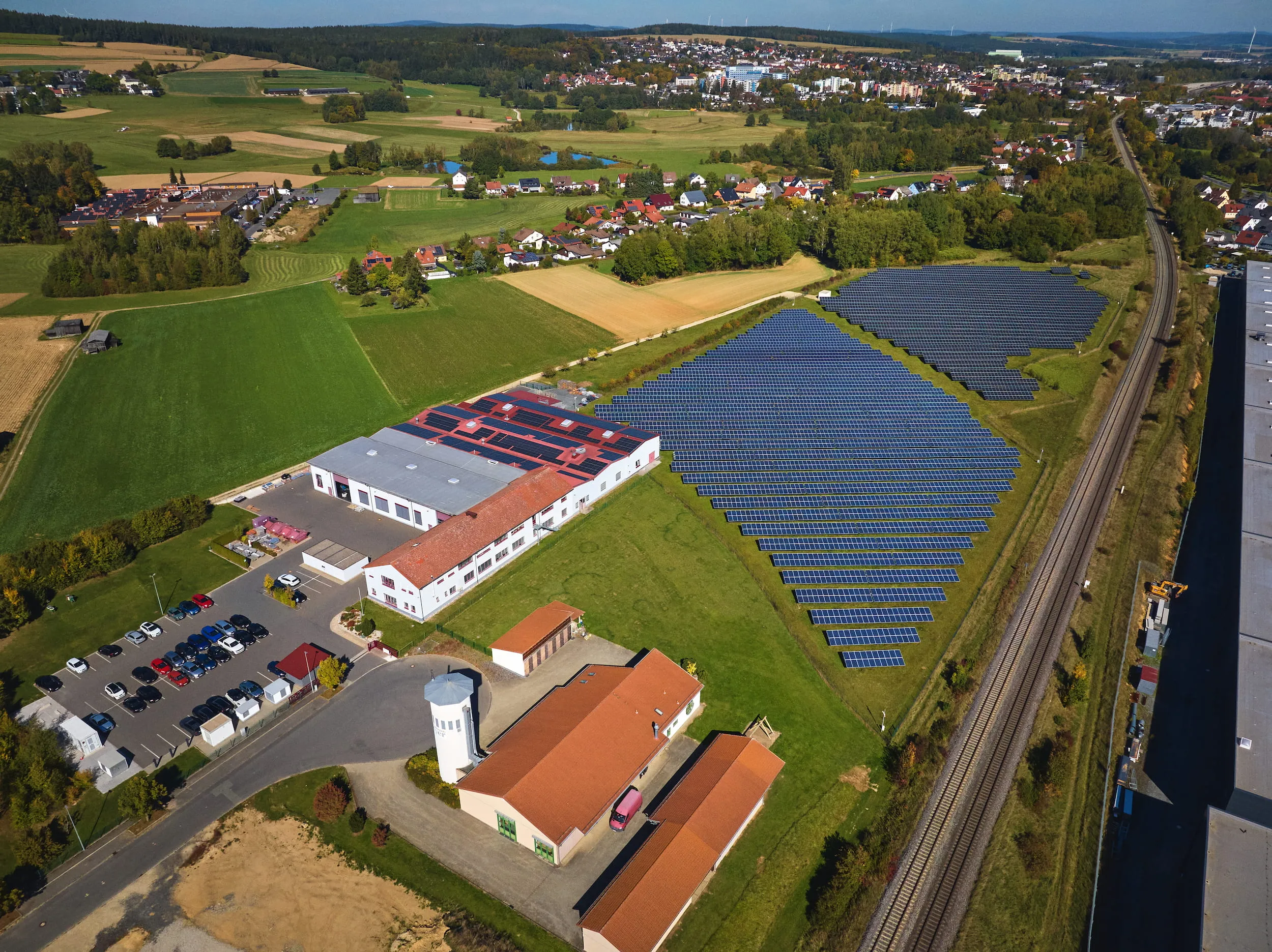 Luftaufnahme des Firmengeländes in Waldershof: weiße Hallen mit Solardach und Parkplatz, daneben ein Solarpark und die Bahnlinie, dahinter Felder und der Ort